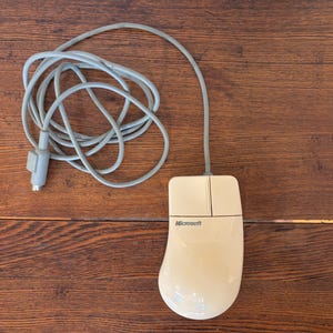 Puede incluir: Un ratón de ordenador Microsoft blanquecino con un cable gris. El ratón tiene dos botones y una rueda de desplazamiento. El logotipo de Microsoft está impreso en la parte superior. El cable está enrollado.