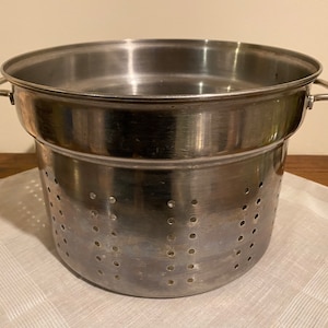Könnte beinhalten: Ein doppelter Edelstahl-Kochtopf mit perforiertem Boden. Der Topf hat zwei Griffe und ist zum Garen von Speisen im Wasserbad konzipiert.
