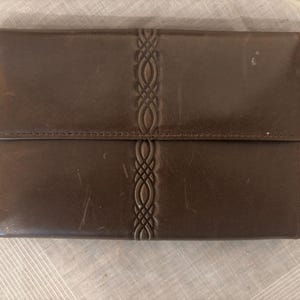 May include: A rectangular brown leather wallet featuring a smooth finish and a vertical central panel embossed with an intricate, repeating woven knot pattern. The wallet has a horizontal flap closure and visible surface scuffs on the leather.