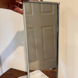 May include: A rectangular mirror with a white frame and a silver reflective surface. The mirror is held by a bolt and nut at the top. The mirror is reflecting a white door.