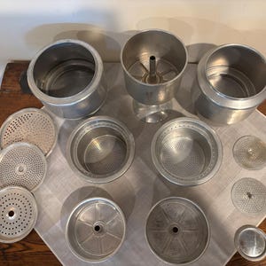 May include: A collection of disassembled silver-toned metal coffee maker components, including three main cylindrical vessels, several circular perforated filter discs, and two deep bowl-shaped filter baskets with fine mesh bases.