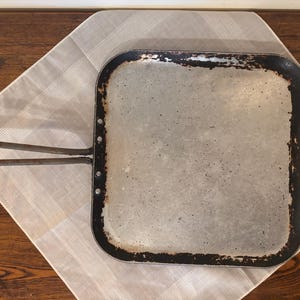 May include: A square metal griddle with a worn, dark finish along the edges and a lighter, speckled center. The cooking surface features a long, thin, looped metal handle attached to one side with three visible rivets, showing signs of use.