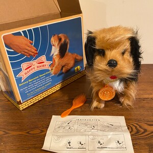 Puede incluir: Un perro de juguete marrón y negro con un collar dorado y una etiqueta redonda que dice "Happy Hairy". El perro funciona con pilas y tiene control de sonido. El perro está sentado sobre una superficie de madera con una caja marrón y un cepillo naranja.