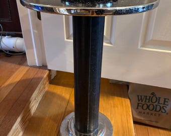 Puede incluir: Un cenicero de pie vintage con una tapa negra y un borde cromado. El soporte tiene una columna negra estriada y una base cromada. Un cenicero cromado se encuentra encima. El soporte está sobre una superficie de madera.