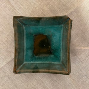 May include: A small, square ceramic dish with a teal glaze and a brown center. The dish has a slightly raised rim.
