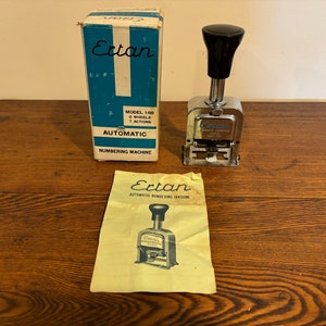 May include: A silver automatic numbering machine with a black handle. The machine is labeled "Ertan" and "Model 150 6 Wheels 7 Actions". The machine is on a wooden surface with a yellow instruction sheet.