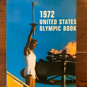 以下が含まれることがあります： 青い背景に「1972 UNITED STATES OLYMPIC BOOK」の白い文字が書かれたヴィンテージ本の表紙。表紙には、青い空を背景に、炎を上げたトーチを持つアスリートが描かれています。