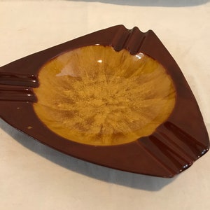 May include: A triangular ceramic ashtray with a brown rim and a yellow-brown center. The ashtray has three grooves on each side for cigarettes.