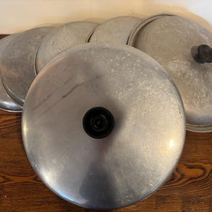 Puede incluir: Un juego de seis tapas de metal de color plateado para ollas y sartenes. Las tapas están apiladas una encima de la otra, con la tapa más grande en la parte inferior y la tapa más pequeña en la parte superior. Las tapas tienen una superficie lisa y brillante.