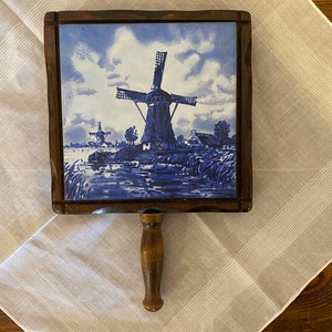 Puede incluir: Una bandeja de servir de madera con una baldosa azul y blanca que representa una escena de molino de viento. La baldosa está enmarcada en madera y la bandeja tiene un asa de madera.