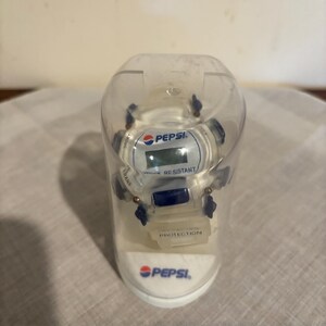 Puede incluir: Un reloj de pulsera digital blanco con detalles en azul dentro de una cúpula de plástico transparente. La esfera muestra el logotipo de Pepsi, el texto SHOCK RESISTANT y la palabra PROTECTION en la correa. La base también tiene el logo de Pepsi.