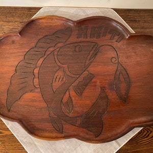 Puede incluir: Una bandeja de servir de madera con un diseño tallado de dos peces. La bandeja es marrón y tiene un borde festoneado. La palabra "HRIITI" está tallada en la parte superior de la bandeja.