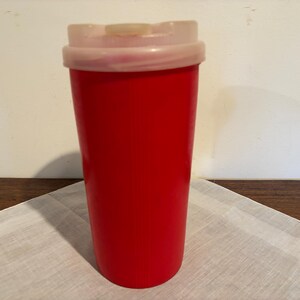 May include: A tall, cylindrical red tumbler with a smooth, solid-colored exterior. The vessel features a translucent, off-white plastic lid that fits securely over the top rim, providing a simple and functional design for holding beverages.