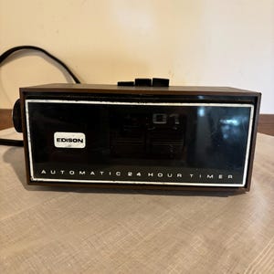 Puede incluir: Un temporizador automático Edison vintage de 24 horas. El temporizador tiene un marco de madera marrón oscuro con una cara negra y texto blanco. La pantalla muestra el número "01". La palabra "EDISON" está impresa en el lado izquierdo de la cara.