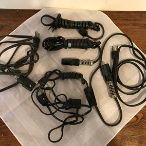 Puede incluir: Cinco cables eléctricos negros con varios enchufes y conectores. Los cables están enrollados y enredados sobre una superficie blanca.