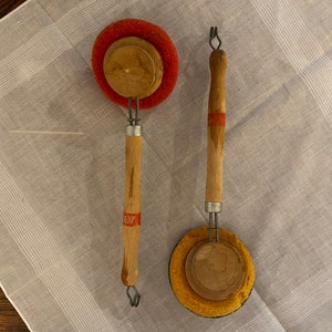 May include: Two vintage dish scrubbers with wooden handles and red and yellow scouring pads. The scrubbers have metal wire loops at the top.