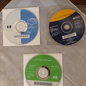 Op de afbeelding: Drie HP printersoftware-cd's worden weergegeven op een lichtgekleurd oppervlak. De cd's zitten in witte papieren hoezen. Eén cd is gelabeld "HP LaserJet 1150/1300/1300n", een andere "HP DeskJet 840C Series" en de derde "HP Deskjet D2500 series".
