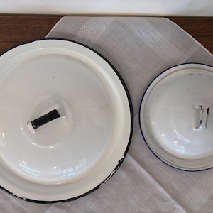 Op de afbeelding: Twee witte emaille deksels met zwarte randen en handvatten. Het grotere deksel heeft een rechthoekig zwart handvat, terwijl het kleinere deksel een gebogen handvat heeft. Beide deksels liggen op een witte doek.