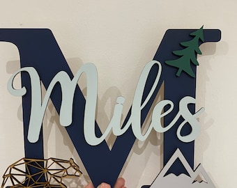May include: A navy blue wooden letter M with the name "Miles" in light blue cursive lettering. The letter is decorated with a gold geometric bear, a green pine tree, and gray mountains.