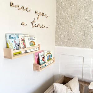 May include: Wooden wall shelves with books on them. The shelves are mounted on a white wall. The text "once upon a time..." is written on the wall in wooden letters.