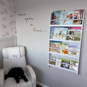 Könnte beinhalten: Ein weißes Bücherregal mit Kinderbüchern ist an einer grauen Wand befestigt. Unterhalb befindet sich ein weißer Relaxsessel mit einem schwarzen Stofftier. An der Wand steht der Text "once upon a time..." und eine gemusterte Tapete.