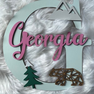 May include: A light blue wooden letter G with the name "Georgia" in pink letters. The letter is decorated with a white mountain range, a green pine tree, and a geometric brown bear.