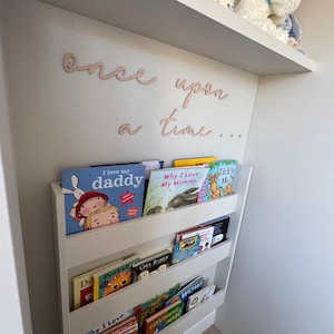 May include: A white bookshelf unit filled with children's books, with the words "once upon a time..." in a cursive font on the wall. A stuffed toy and a small book are on the shelf above.
