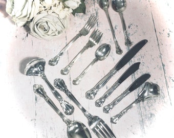 May include: A set of silver flatware, including forks, spoons, and knives, arranged on a white wooden surface. The flatware has an ornate design and is in excellent condition.