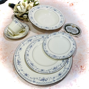 May include: A set of four white ceramic plates with a blue floral pattern and a silver rim. The plates are arranged in a stack, with the largest plate on the bottom and the smallest plate on top. The plates are made by Minton.