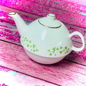 Op de afbeelding: Witte theepot met een groen klaverblad ontwerp. De theepot heeft een gouden rand rond de deksel en de tuit.
