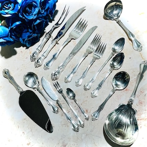 May include: A collection of silver-plated serving utensils, including a serving spoon, a ladle, a cake server, and several forks. The utensils are arranged on a white surface.