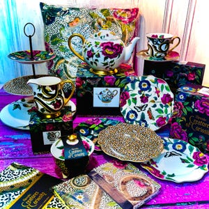 May include: A Spode Creatures of Curiosity tea set featuring a teapot, creamer, sugar bowl and two plates. The set has a black and white leopard print design with gold accents. The teapot features a floral design with pink flowers. The set is displayed against a pink and purple background.