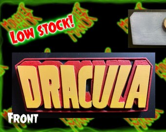 May include: A 3D wooden sign with the word "Dracula" in raised yellow letters with red outlines. The sign is on a black background. The back of the sign is shown with a magnet attached. The sign is $15.00.