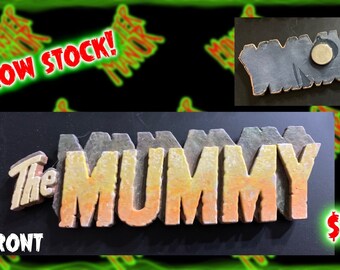 May include: A 3D wooden sign with the words "The Mummy" in a yellow, orange, and brown gradient. The sign has a distressed, aged look. The back of the sign is plain gray. The sign is $15.00.