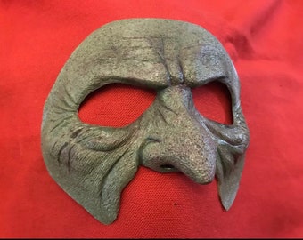 May include: A green, textured, moulded plastic mask with a large, bulbous nose and eye holes. The mask is designed to resemble a grotesque, aged face.