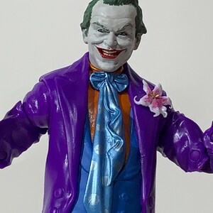 May include: A detailed action figure of the Joker, featuring a purple suit, orange shirt, and blue tie. The figure has green hair, white face paint, and a red smile. A small flower is pinned to the lapel. The figure is a collectible item.