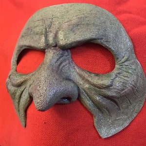 May include: A green, textured, and detailed mask with a large, prominent nose and eye holes. The mask is made of a material that resembles leather or rubber.