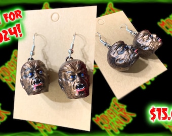 May include: A pair of silver dangle earrings with bronze werewolf head charms. The earrings are displayed on a brown card with the text "NEW FOR 2024!" and "$15.00".