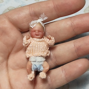 Puede incluir: Una muñeca en miniatura de tez pálida, que lleva un suéter de punto color melocotón claro, una diadema de encaje blanco y un pañal pequeño. La muñeca tiene los ojos cerrados y una expresión neutra, en una escala pequeña para la palma.