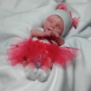 Puede incluir: Una pequeña muñeca bebé vestida con un gorro de punto blanco con lazos rojos, un body blanco y un tutú de tul rojo. La muñeca está acostada sobre una tela blanca, con los ojos cerrados y las manos cerca de la cara.