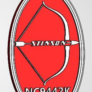 May include: Oval-shaped pendant with a red background and a white bow and arrow design. The word "STINSON" is written across the arrow. The pendant has a white border and a loop at the top. The text "NC9442K" is at the bottom.
