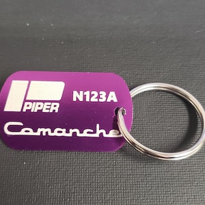 May include: A purple Piper Comanche keychain with a silver key ring. The keychain has white text that reads "PIPER", "N123A", and "Comanche". The keychain is rectangular with rounded corners.