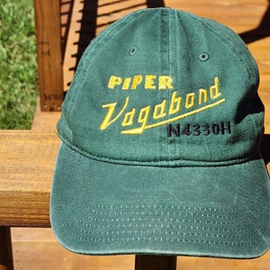 May include: Green baseball cap with yellow embroidered text that reads "PIPER Vagabond N4330H".