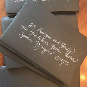 May include: A stack of dark gray envelopes with white calligraphy. The envelopes display addresses, including names, street addresses, and zip codes. The addresses are written in an elegant, cursive font.