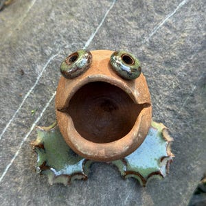 May include: A small, round, unglazed terracotta-colored ceramic frog figurine with a wide, open mouth. The frog features two raised, glossy, dark green glazed eyes and two flat, scalloped, light green glazed feet extending from the base.
