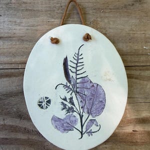 May include: An oval-shaped white ceramic wall plaque featuring a purple botanical leaf and fern imprint. The plaque has two small holes at the top with a brown leather cord threaded through for hanging, and a small circular stamp on the left side.