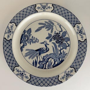 May include: A round ceramic plate featuring a blue and white transferware pattern. The center depicts a bird standing among stylized flowers and foliage. The wide rim is decorated with a geometric lattice border and floral motifs.