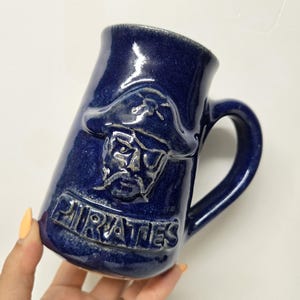 May include: A navy blue ceramic mug with a handle. The mug features a raised design of a pirate with an eye patch and the word "PIRATES" below. The mug is held by a hand.