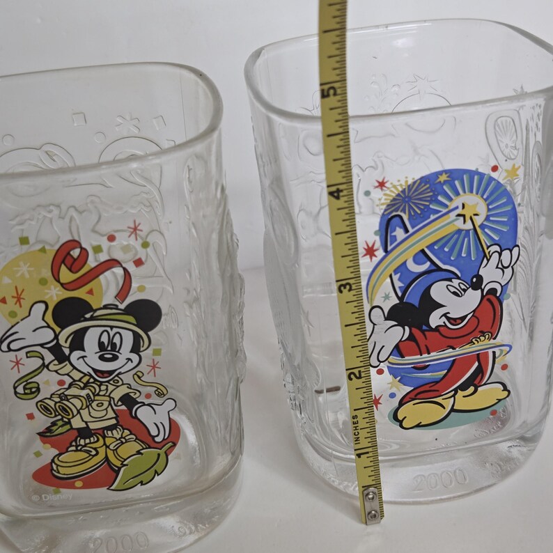 May include: Two clear Disney glasses with Mickey Mouse graphics. One glass features Mickey in safari attire, the other shows Mickey as a sorcerer. The glasses are approximately 5 inches tall. The year "2000" is printed on the bottom of each glass.