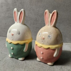 May include: Two ceramic bunny figurines, one with a green and white outfit and the other with a pink and white outfit. Both bunnies have yellow collars and pink ears.